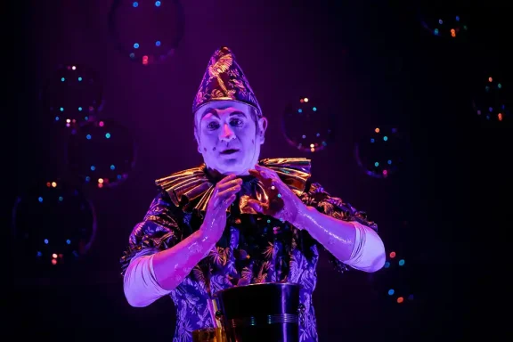 Seifenblasenclown Stephan Masur in poetischer Seifenblasenshow Ein Performer in buntem Kostüm steht unter bunten Lichtern und Blasen.