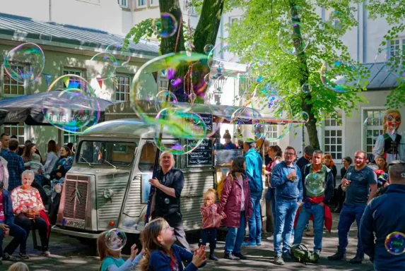 Riesenseifenblasen Walkact Hennschen Theaterfest Menschenmenge auf einer Straßenfest, umgeben von Seifenblasen und einem alten Fahrzeug.