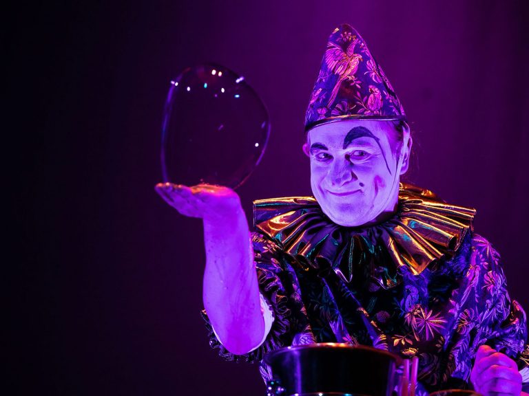 Mann gestaltet große Seifenblasen auf der Bühne, umgeben von buntem Licht.
