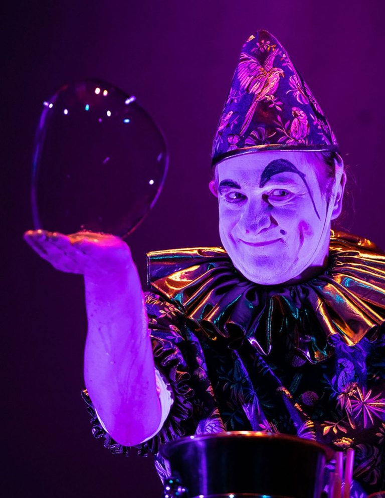 Handseifenblase Stephan Masur als Clown Clown in bunting holds eine schimmernde Seifenblase vor einem lila Hintergrund.