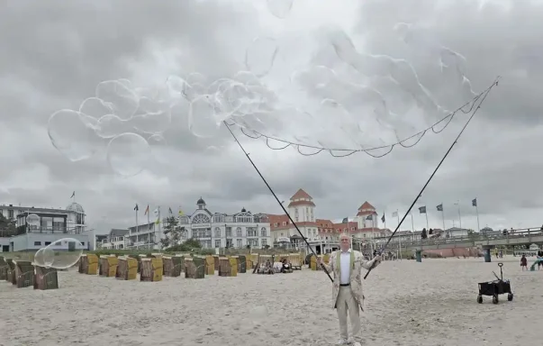 Seifenblasensturm am Strand Ein Mann steht am Strand mit zwei langen Stäben, im Hintergrund sind Strandkörbe und Wolken.