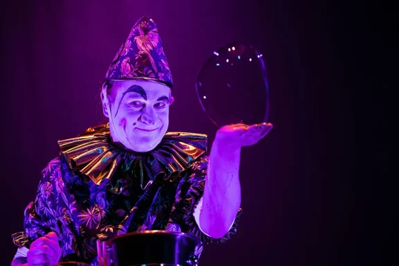 Clown mit glitzerndem Kostüm hält eine Seifenblase vor einem lila Hintergrund.