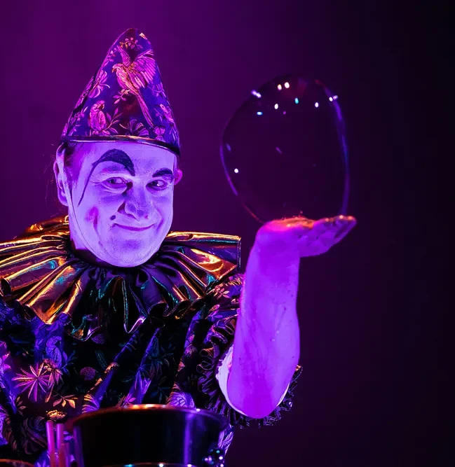 arneval für Kinder in Köln: Seifenblasen-Künstler Stephan Masur mit magischen Riesenblasen clown in schillerndem Kostüm hält durchsichtigen Ballon vor sich.