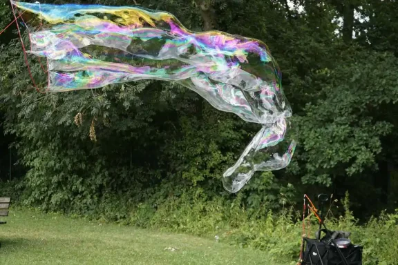 Riesenseifenblase Große bunte Seifenblase schwebt über einer Wiese neben einem schwarzen Koffer.