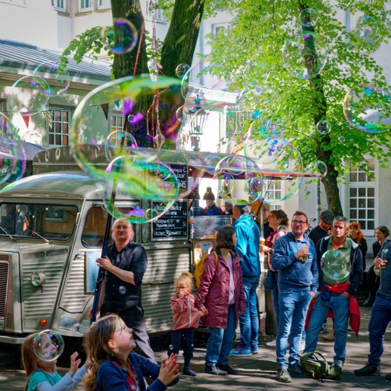Seifenblasen Walk Act Seifenblasenkünstler Masur Menschenfeier mit Seifenblasen und einem Food-Truck, umgeben von Bäumen.