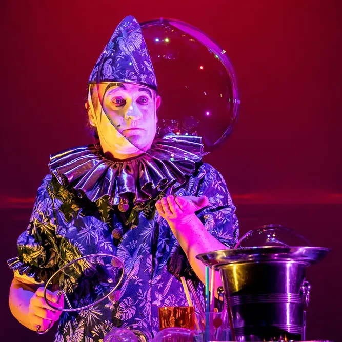 Ein Clown in bunter Kleidung mit einem riesigen Seifenblasen-Ring auf der Bühne.