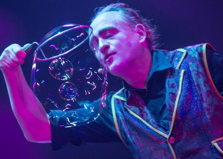 Mann mit Getränk an einem Tisch, neben einer Feuerquelle auf der Bühne.