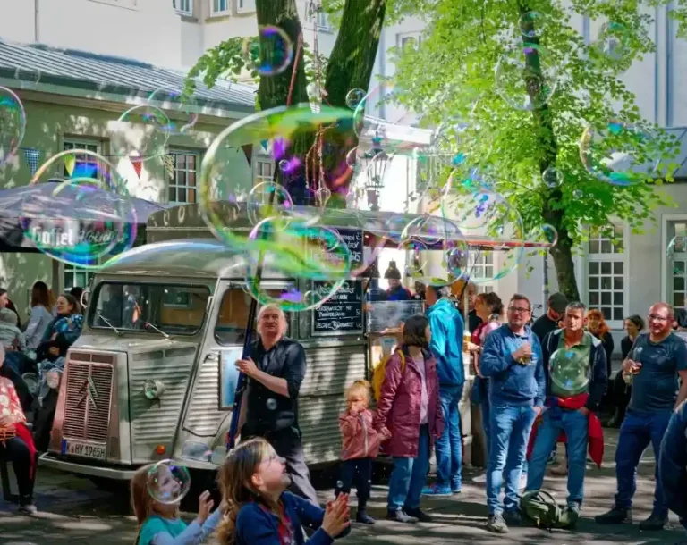 Seifenblasenwalkact auf Gala Straße mit Menschen, die Seifenblasen genießen, und einem alten Fahrzeug im Hintergrund.
