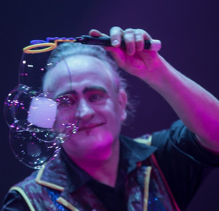 Seifenblasenkünstler Stephan Masur Seifenblasenshow Berlin Ein Zauberer jongliert mit einer Seifenblase vor buntem, schimmerndem Hintergrund.