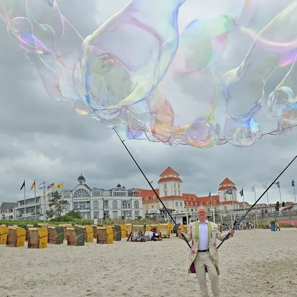 Seifenblasenstürme in ienem partizipativen Seifenblasen Walk Act Ein Mann in einem Anzug hält große Seifenblasen am Strand mit Haustür.