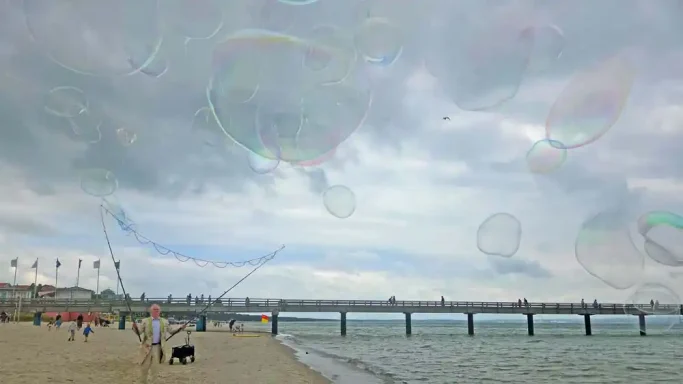 Riesenseifenblasen Walkact am Strand Stephan Masur Strand mit Menschen, Meer im Hintergrund und schimmernden Seifenblasen im Himmel.