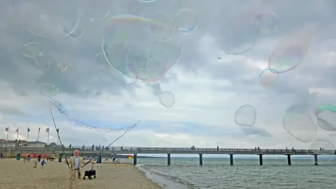 Seifenblasenartist Stephan Masur mit Riesenseifenblasen Menschen am Strand unter einem bewölkten Himmel, mit Seifenblasen im Vordergrund.