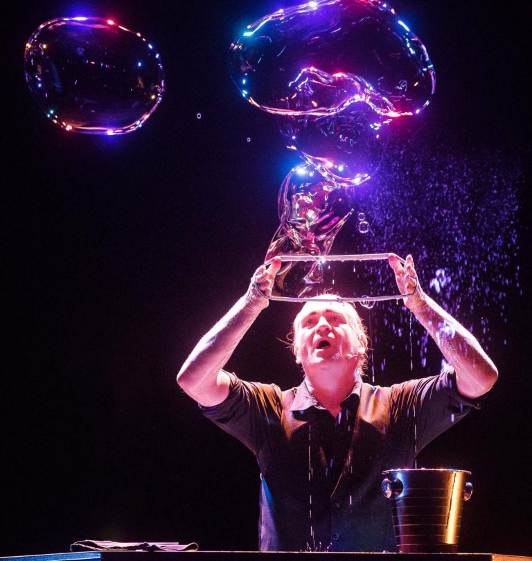Mann gestaltet große Seifenblasen auf der Bühne, umgeben von buntem Licht.