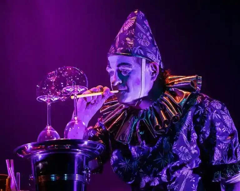 Kleine Events, maximale Qualität – Seifenblasenshow mit dramaturgischer Tiefe Clown mit buntem Kostüm bläst Seifenblasen auf einer Bühne in lila Beleuchtung.