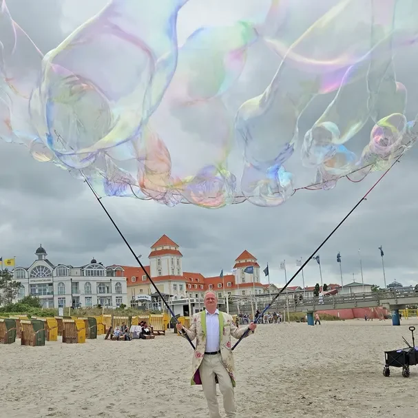 Fragile Schönheit – Das Authentische an der Vergänglichkeit von Seifenblasen Mann mit großen Seifenblasen auf dem Strand, im Hintergrund Bauten und Wolken.