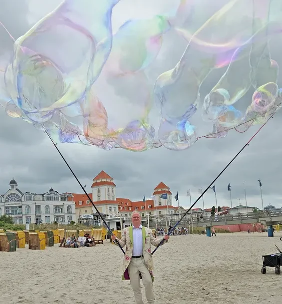 sensorische Seifenblasen auf Festival Mann hält große Seifenblasen über einem Strand mit einem historischen Gebäude im Hintergrund.