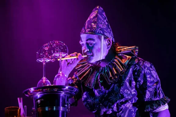 Ein Clown mit buntem Kostüm bläst Seifenblasen vor einer lila Hintergrundbeleuchtung.