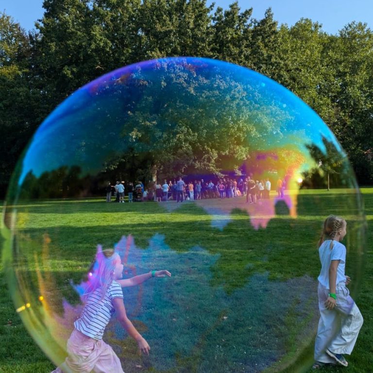 Ein Kind spielt mit Seifenblasen im Park, umgeben von anderen Personen und Bäumen.