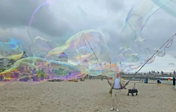 Seifenblasenwalkact am Strand Stephan Masur Mensch mit Hund am Strand, der große Seifenblasen erzeugt. Wolkiger Himmel im Hintergrund.
