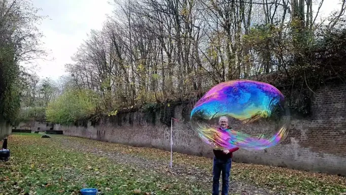 Bllick durch Riesenseifenblasen Person im Park hält eine große, bunte Seifenblase vor einer Baumreihe.