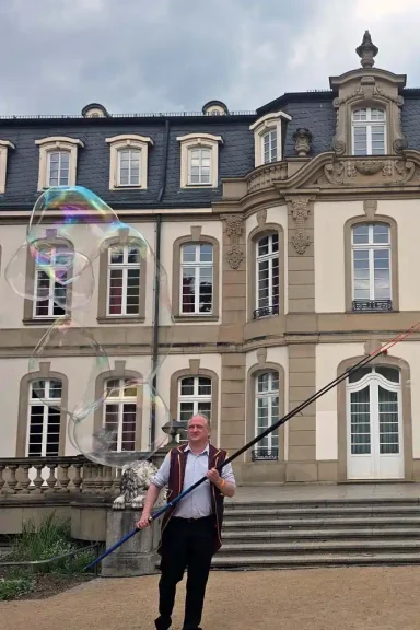 Riesenseifenblasden Büsing Palais Ein Mann mit einem langen Stock bläst Seifenblasen vor einem historischen Gebäude.